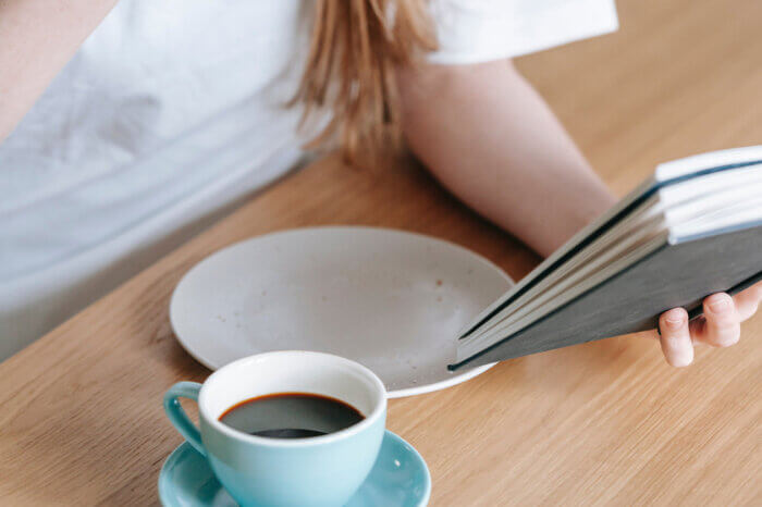 Eine Person sitzt mit einem Buch an einem Tisch, auf dem eine Kaffeetasse steht.